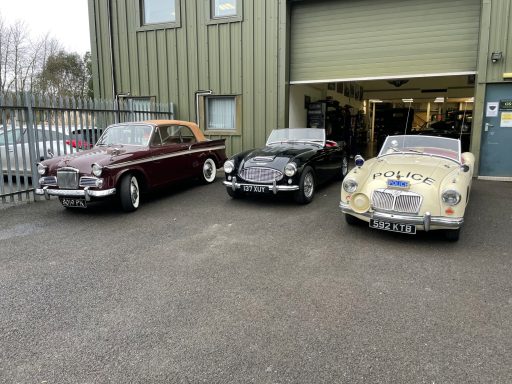 Three classic cars parked outside Chichester Classics Ltd, showcasing vintage styles and colours.